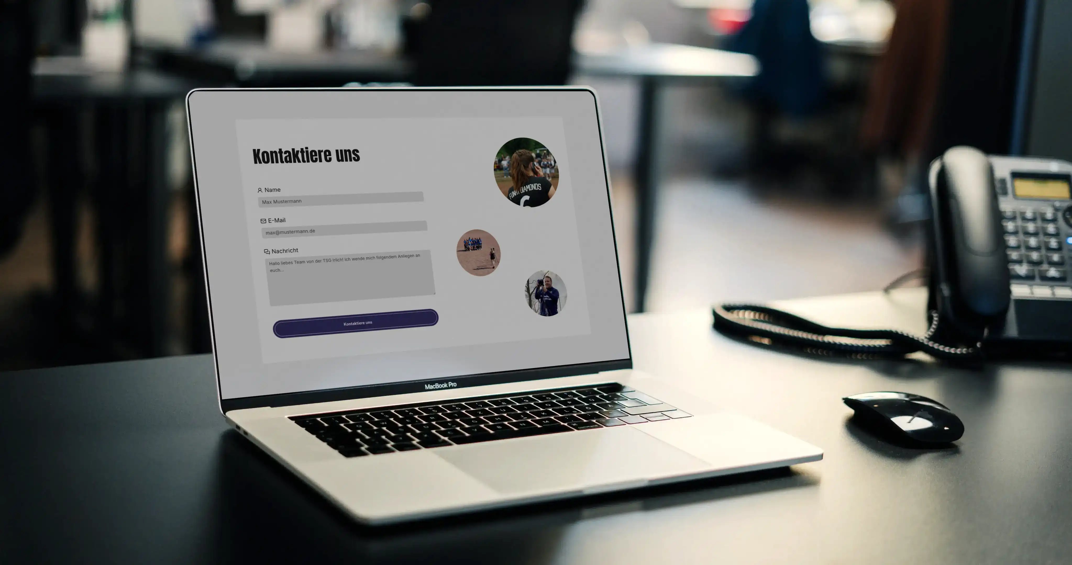 Das Bild zeigt einen modernen Arbeitsplatz. Im Vordergrund steht ein MacBook Pro mit einem ausgeschalteten Bildschirm auf einem schwarzen Schreibtisch. Rechts daneben befindet sich ein Festnetztelefon und eine kabellose Maus. Im Hintergrund ist ein Büro mit unscharfen Personen und Möbeln erkennbar. Die Szene ist gut ausgeleuchtet und vermittelt eine professionelle Arbeitsatmosphäre.